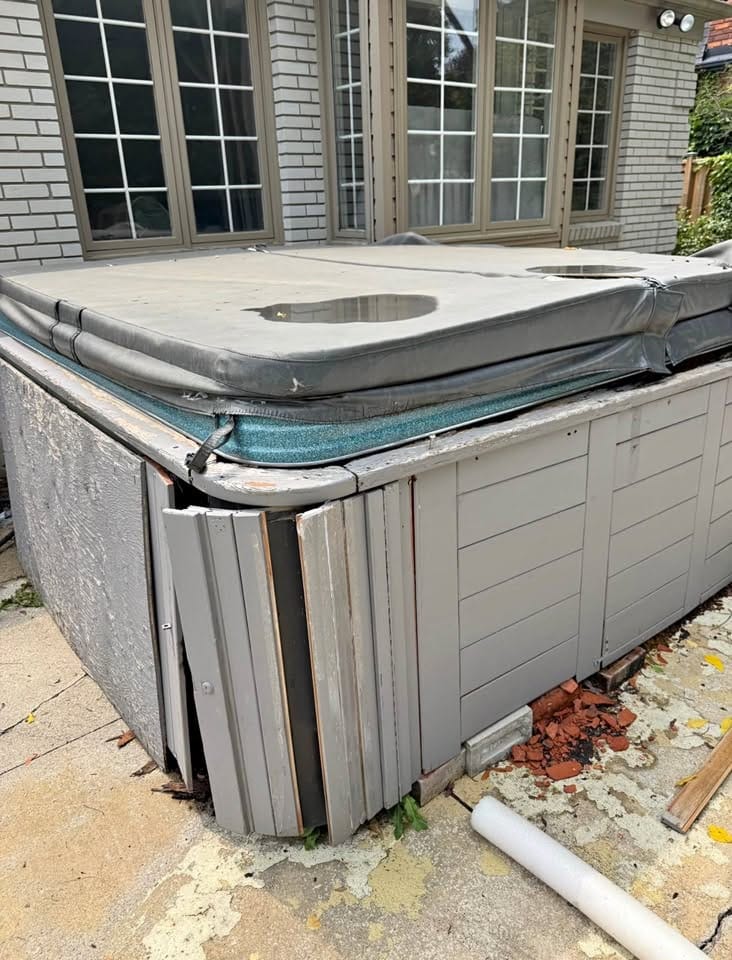 A four-person hot tub beside a brick house with its grey skirting panels pulled away, ready for removal