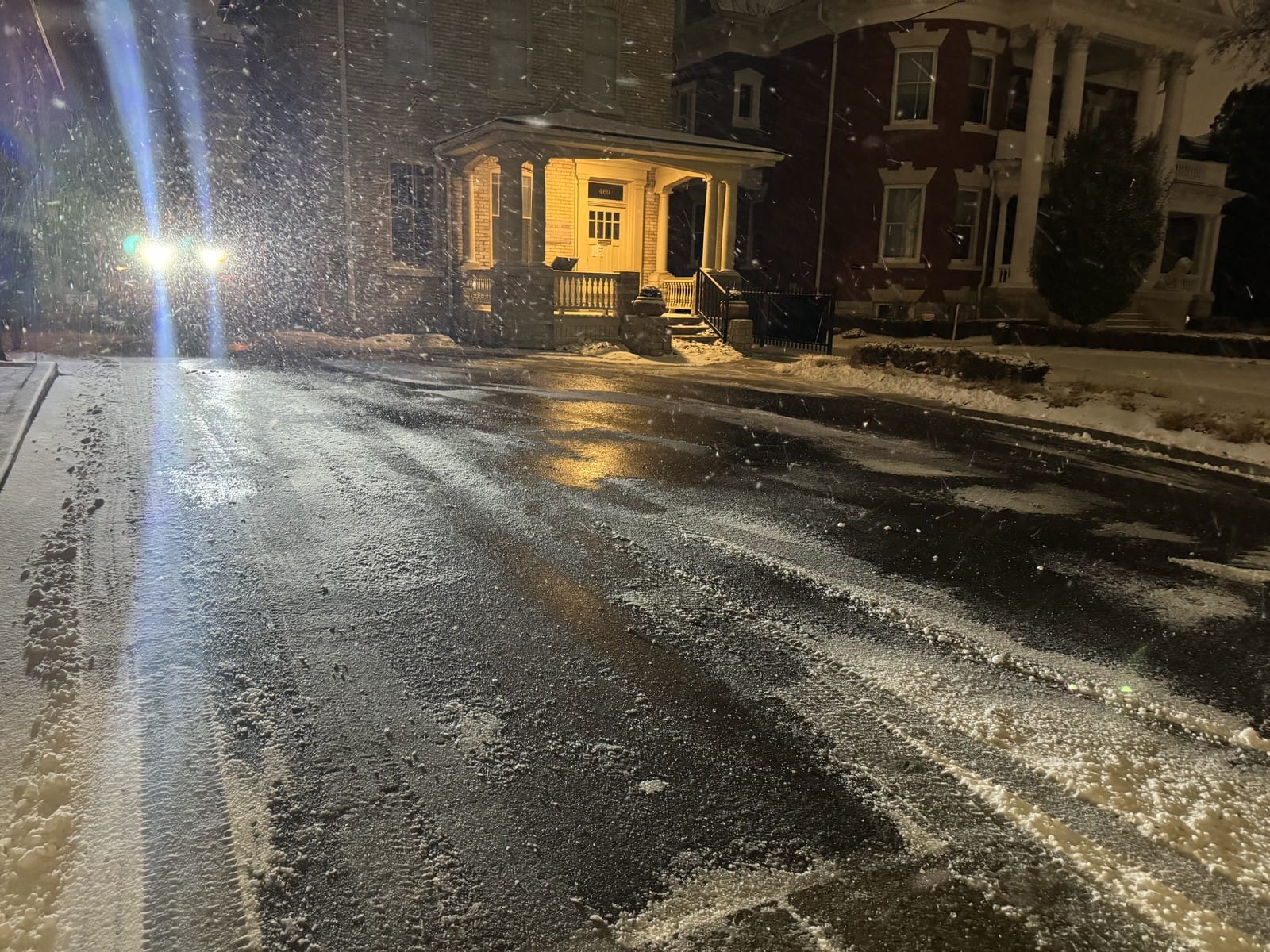 Pavement plowed and salted mid-snowfall at night, wet black surface under streetlight as snow keeps falling