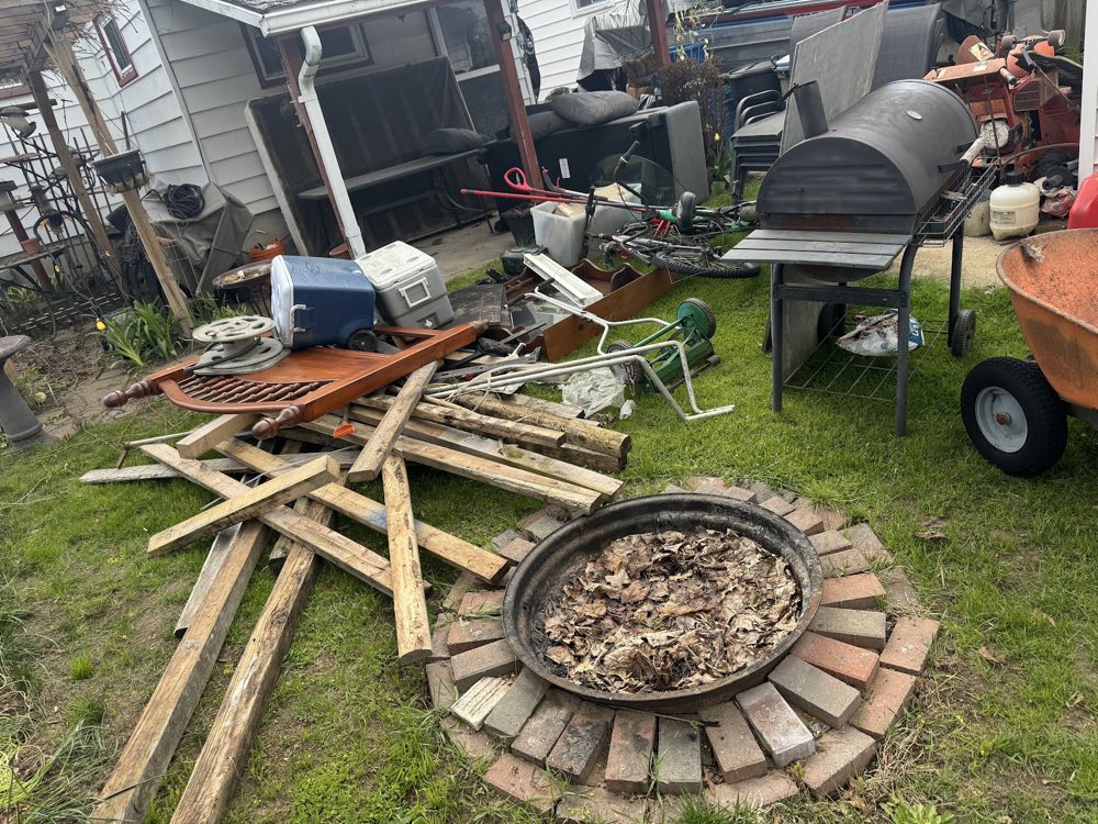 A backyard packed with old lumber, a rusted BBQ, scrap metal and a fire pit being cleared out